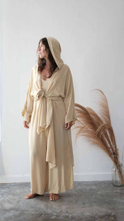 Woman wearing a beige hooded dress standing against a white wall with dried pampas grass.