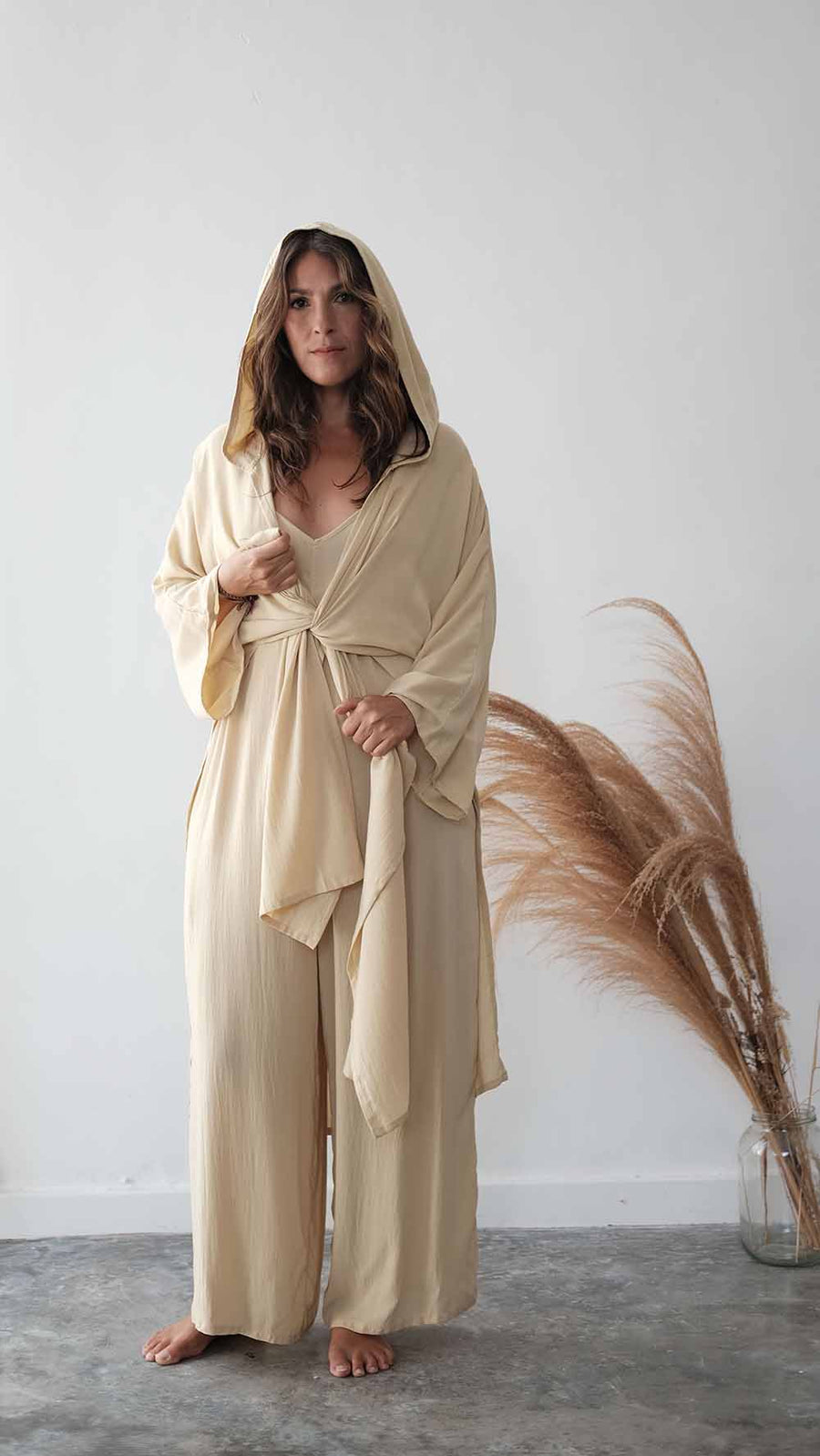 Woman wearing a beige robe standing against a white wall with dried plants.