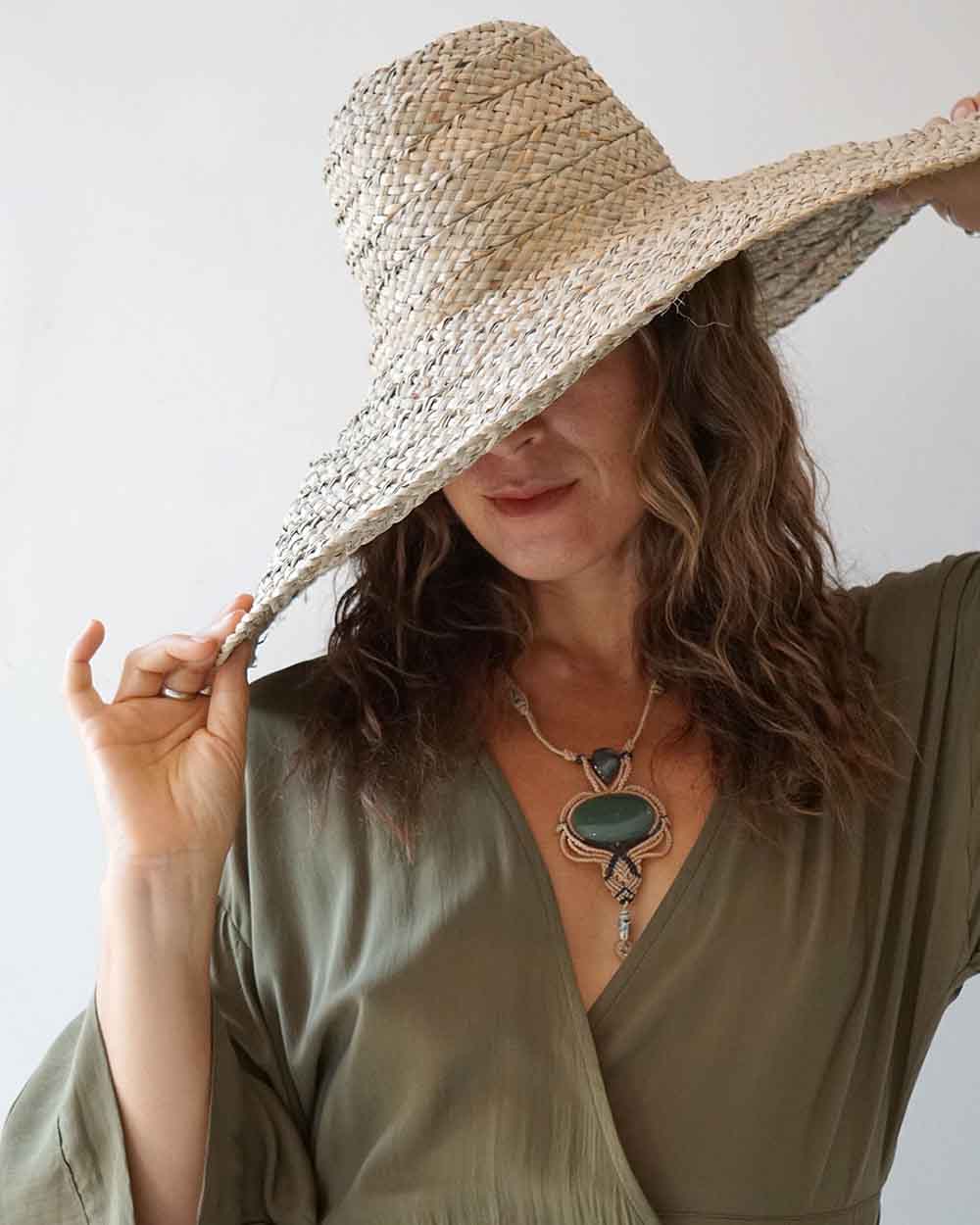 Woman wearing a wide-brimmed straw hat and a green dress with a necklace against a plain background