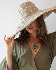 Woman wearing a wide-brimmed straw hat and a green dress with a necklace against a plain background