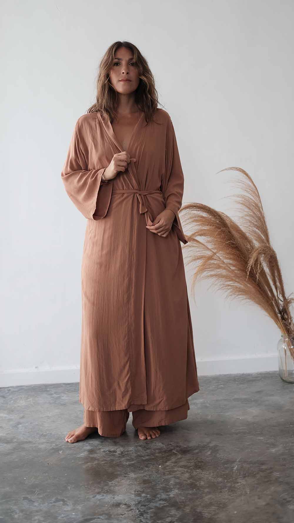 Woman wearing a long brown robe standing against a white wall with dried pampas grass.