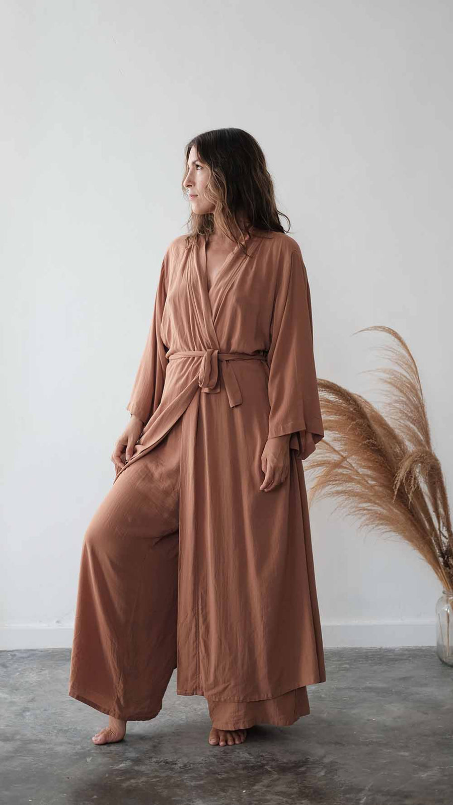 Woman wearing a brown dress standing against a white wall with dried pampas grass.