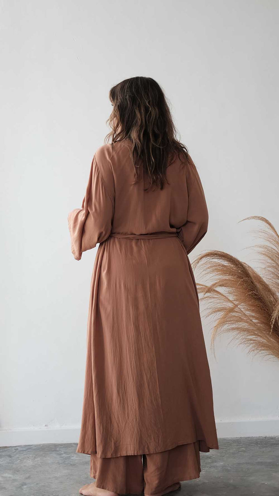Woman wearing a long brown dress standing against a white wall with decorative elements.