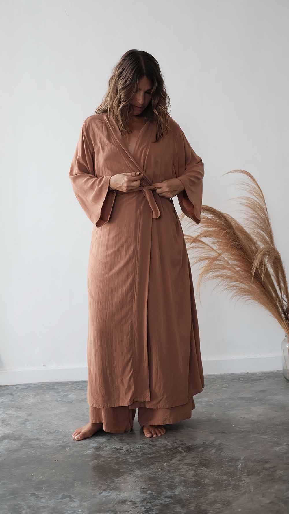 Woman wearing a long brown dress standing against a white wall with a plant to the side.