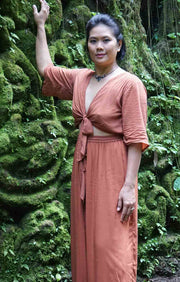Woman in a rust-colored outfit standing against a moss-covered stone wall in a forest setting.
