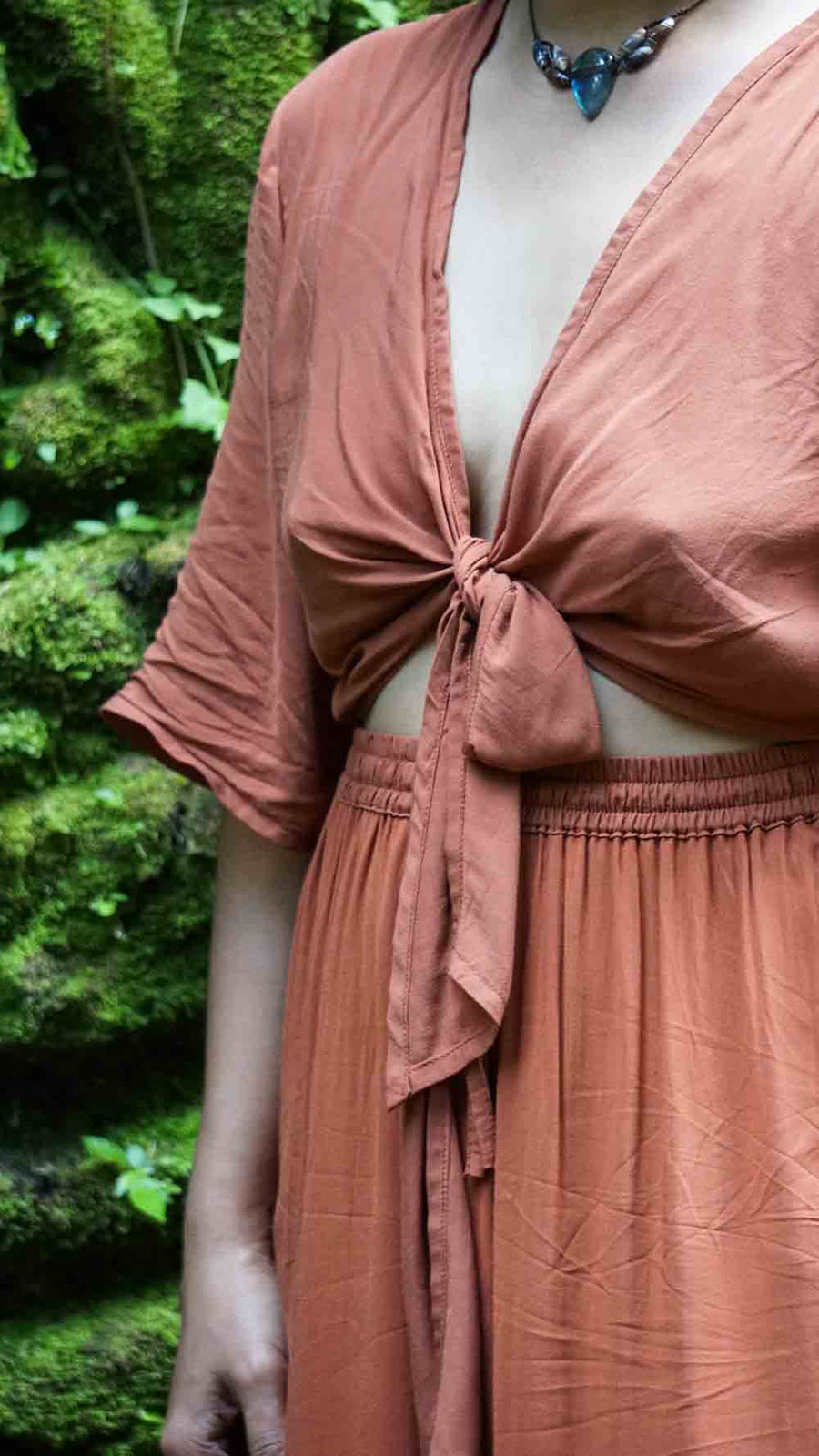 Person wearing a rust-colored dress with a tie detail in front of a green, natural background.