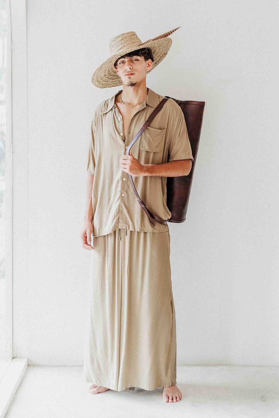 Man wearing a beige outfit with a wide-brimmed hat and carrying a brown bag against a white background