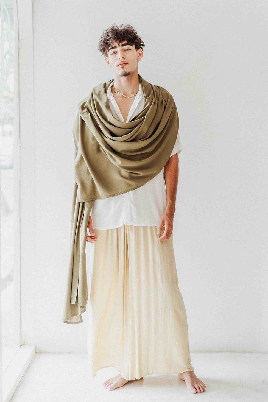 Man wearing a large green shawl over a white shirt and beige pants on a white background