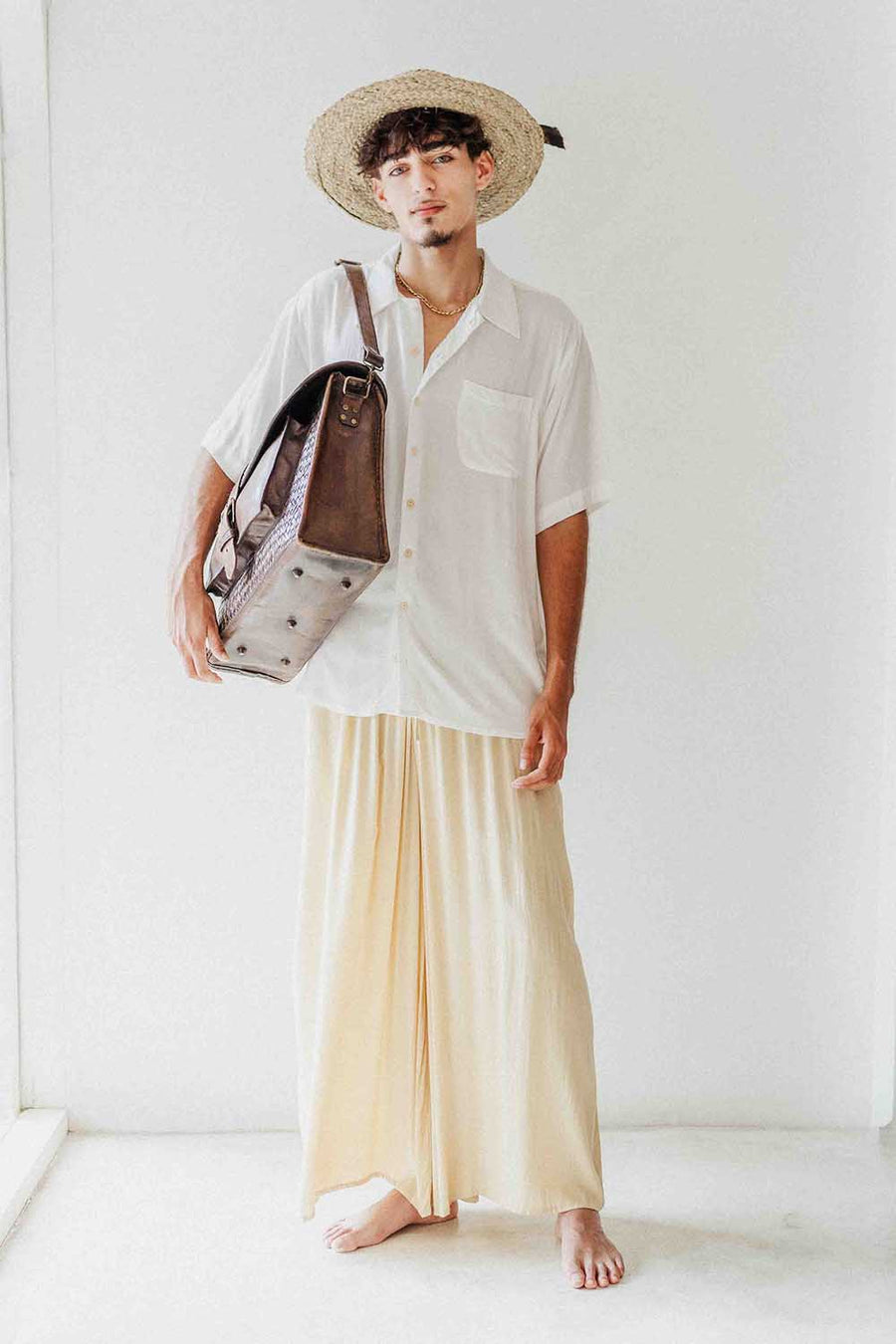 Man wearing a white shirt, beige pants, and a straw hat holding a bag against a white background